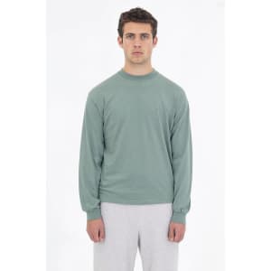 A long sleeve crewneck tee in a soft green hue, worn with light grey bottoms. The shirt features a logo on it.