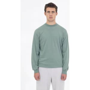 A long sleeve crewneck tee in a soft green colour, featured on a model wearing grey pants. The shirt has a logo.