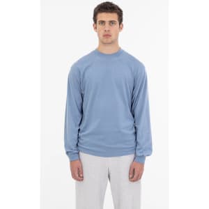 A long sleeve crewneck tee in a soft blue colour, worn by a model with grey pants. It features a small logo.