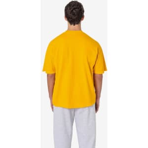A man stands with his back to the camera, wearing a bright yellow short sleeve pocket tee and grey sweatpants.