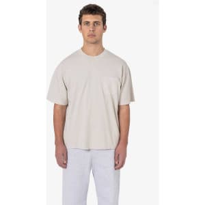 A model wears a beige short sleeve garment-dyed pocket tee paired with grey trousers. The shirt features a pocket.