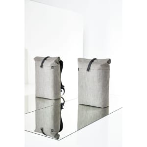 A grey roller backpack with a structured design, featuring a top flap and a logo. Two bags are displayed on a reflective surface.