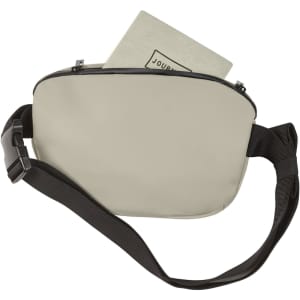 A beige bum bag with a black strap, featuring a zip closure and a journal inside. There is a logo on the bag.