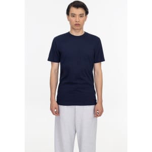 A man stands wearing a navy short sleeve fine jersey crew tee and grey sweatpants. The shirt features a logo.