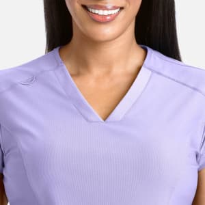 A woman smiles while wearing a lavender banded V-neck top with a logo.