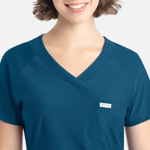 A close-up of a woman wearing a teal Embrace Luna Tuckable Y-Neck Top with a small pocket and branding.
