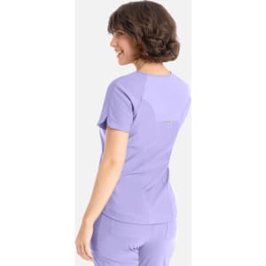 A woman in a lilac Y-neck top is smiling, showcasing the back view of the shirt. The top has a logo.