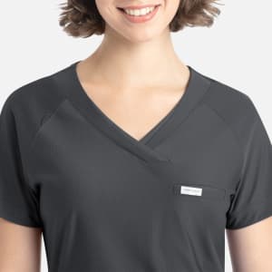 A dark grey Y-neck top featuring a tucked design and a small pocket, modelled by a smiling person with short hair.