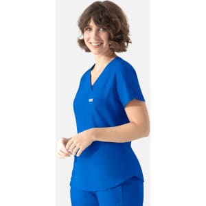 A woman models a blue scrub top with a V-neck and a small pocket. It features a branding logo.