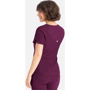 A woman is wearing a purple Embrace Luna Tuckable Y-Neck Top, viewed from the back. The top features a logo.