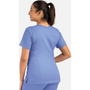 A woman stands with her back to the camera, wearing a light blue double V-neck top. The top features short sleeves and has a logo.
