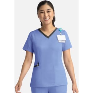 A nurse in a blue scrub top with a contrast V-neck and pocket details, wearing a badge and a smile.