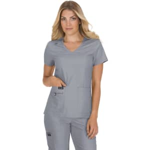 A woman in a grey scrubs top with pockets, posing with hand in pocket. The top has a logo on it.