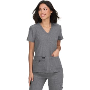 A woman models a grey v-neck top with two front pockets, part of a matching scrubs set. The outfit includes a branding logo.