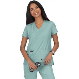 A woman wearing a teal scrub top, posing with a stethoscope. The top features two pockets and branding on the left side.