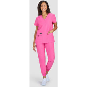 A woman stands in a pink scrub top and matching pants, featuring pockets and a logo on the top.