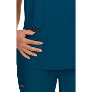 A teal top with a patterned pocket, featuring a hand resting inside the pocket. The garment has a branding logo.