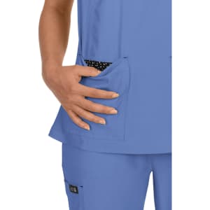 A scrub top in light blue, featuring two front pockets with a contrasting black and white polka dot lining.