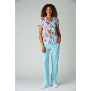 A stylish medical top featuring a colourful floral pattern, paired with light blue scrubs. The model stands smiling.