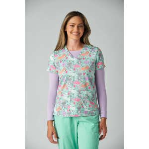 A woman models a colourful scrub top with a floral pattern, featuring shades of pink and green, layered over a long-sleeve top.