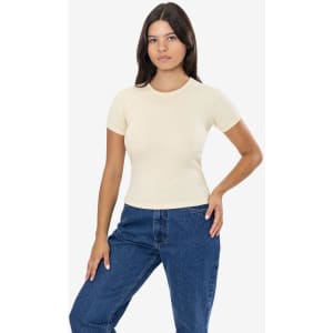 A woman wearing a light beige short-sleeved baby rib tee and blue jeans, standing confidently. The tee has a logo.