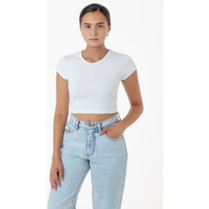 A woman stands wearing a white cap sleeve crop top paired with light blue jeans. The outfit features a logo.