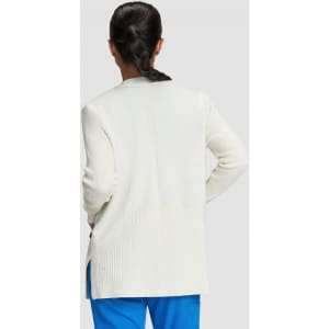 A woman stands with her back to the camera, wearing a white Kori Sweater and blue pants.