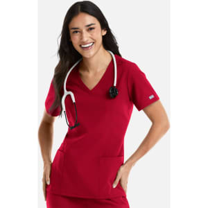 A woman wearing a red double V-neck top with a stethoscope around her neck, smiling and standing with hands on her hips.