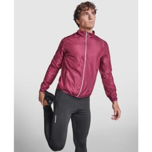 A model wearing a burgundy lightweight windbreaker, paired with black athletic pants, in a stretching pose.