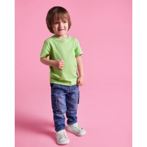 A young child wearing a light green t-shirt and blue jeans, standing against a pink background.