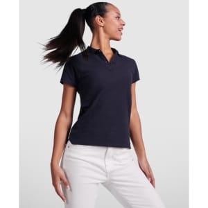 A navy women's polo shirt with a collar, displayed by a model wearing white pants. The shirt features a logo.