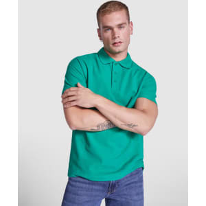 A green Austral Polo Shirt worn by a model with short hair, standing with arms crossed. The shirt has a collar and features a logo.
