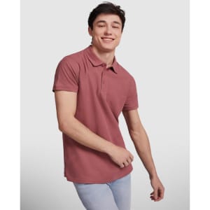 A young man in a burgundy polo shirt is smiling while posing against a neutral background. The shirt features a logo.