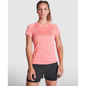 A woman stands in a coral pink sports T-shirt paired with black shorts, featuring a simple logo on the shirt.
