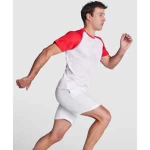 A man in a red and white T-shirt runs, wearing white shorts. The shirt features a logo on the front.