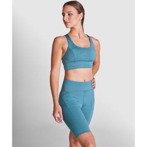 A woman poses in a teal sports cropped tank top and matching high-waisted shorts, featuring a logo.