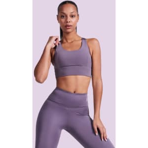 A woman poses in a purple cropped tank top and matching leggings, with a simple background. The top has a logo.