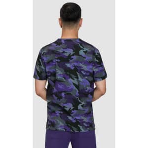 A camouflage print t-shirt in shades of purple, green, and black, viewed from the back. The model is wearing purple pants.