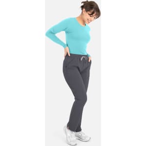 A woman wearing a turquoise long-sleeve tee and grey pants, smiling and looking down at her outfit.