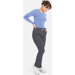 A woman in a light blue long-sleeve top and grey pants, smiling and standing with her hands in her pockets.