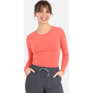 A woman in a coral long-sleeve top, smiling while wearing grey pants. The top features a logo.