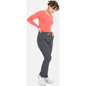 A woman wears a coral long-sleeve underscrub tee with grey pants, smiling and posing. The outfit features pockets.