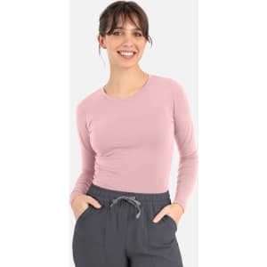 A woman wears a pink long-sleeve underscrub tee paired with grey pants and has her hands in her pockets.