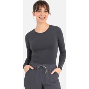 A long-sleeve women's underscrub tee in grey, worn by a model with hands in pockets, featuring a logo.