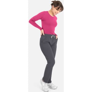 A woman wears a pink long-sleeve underscrub tee with grey pants and white sneakers, smiling while looking down.