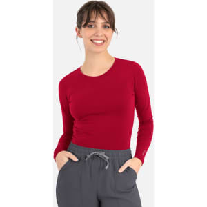 A fitted long-sleeve underscrub tee in red, worn with grey pants, featuring a logo on the sleeve.