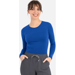 A woman wearing a long-sleeve, bright blue underscrub tee with a fitted style and grey pants. The tee has a logo.