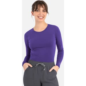 A purple long-sleeve underscrub tee worn by a smiling woman, paired with grey pants. The tee features a logo.