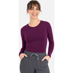 A woman smiles while wearing a long-sleeve underscrub tee in deep purple, paired with grey pants.
