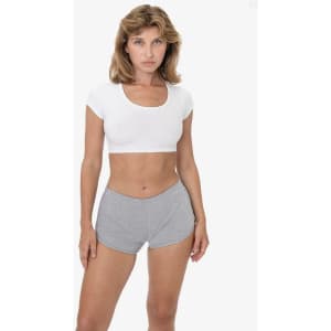 A woman in a fitted white top and grey sports shorts, standing against a plain background. The shorts have a logo.